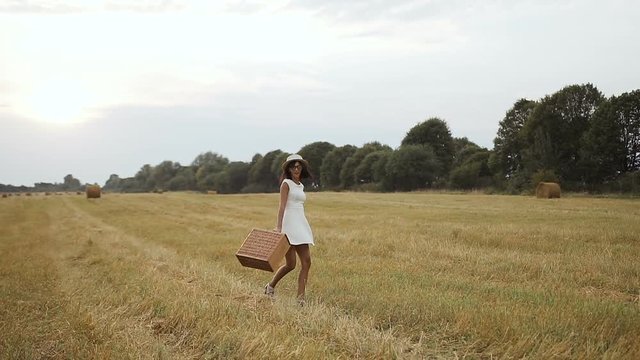 A young girl wearing a white dress with a straw hat and a suitcase. Straw and straw bales. Sunset