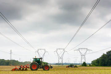 High-voltage lines and transmission towers with tractors plowing a field in the foreground, under a heavy sky.