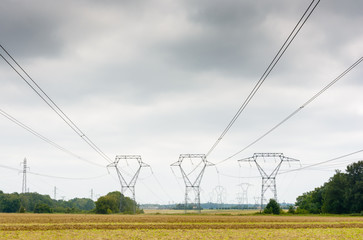 Dozens of transmission towers with high-voltage lines passing over a plowed field under a heavy sky.
