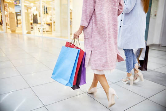 Low section portrait of two beautiful young women wearing coats and high heels walking in shopping mall holding paper bags with purchases