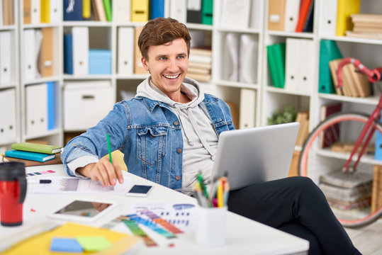 Portrait of smiling young man dressed in casual clothes using laptop while working in modern office