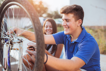 Teenager boy repair tire on bicycle , female friend sitting next to him, summer outdoor photo