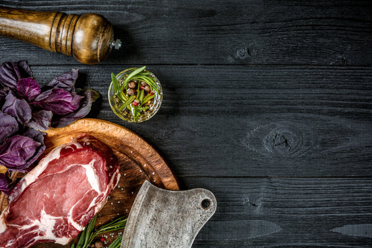 Fresh raw beef with basil and a sprig of rosemary with ax for meat on black wooden background. Top view