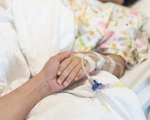 Obraz premium Nursing caretaker concept with kid patient sleeping in bed with family caregiver hand support in blur medical hospital background (focus on hand)