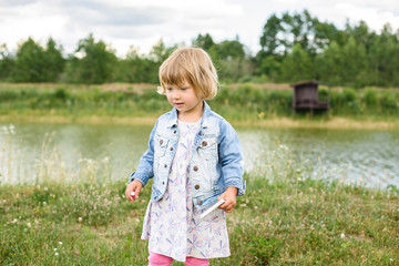 Sad lovely little baby girl with a spinner toy in the hands walks around a small lake outdoors in...