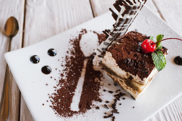 Tiramisu on white square cut plate with decoration from chocolate, cherry and mint. Sweet dessert serving in restaurant on wooden table