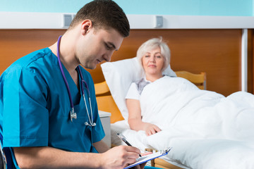 Handsome young male doctor in uniform with phonendoscope on his neck writing down complaints of patient