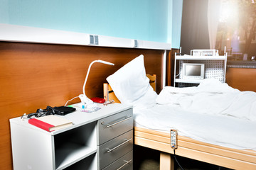 A bedside table with a lot of tablets, a lamp and glasses stands near the empty hospital bed in the hospital ward with medical equipment