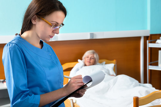 Attractive Nurse In Uniform Wearing A Mask And Glasses Making Some Notes In Report At Clipboard