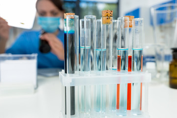 Close up of test tubes with different liquidity, female scientist in uniform wearing a mask on background