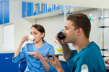 Two young scientists in uniform drinking a coffee after some research