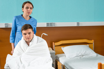 Attractive young nurse pulling wheelchair with ill patient covered with quilt in hospital ward