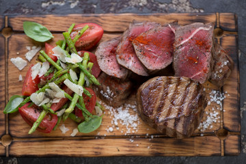 Above view of a wooden serving tray with whole and sliced grilled beef medallion steaks served with asparagus and tomato salad, closeup