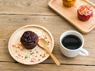 chocolate cupcake put on a spherical wooden plate. Beside of cupcake have Vintage alarm clock and white coffee mug.In the background have yellow cupcake and Red cupcakes.