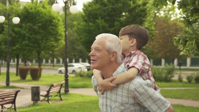 Handsome senior man holding his grandson on his back outdoors. Attractive little boy riding his grandpa on walkway in the park. Brunette caucasian kid pointing his hand forward