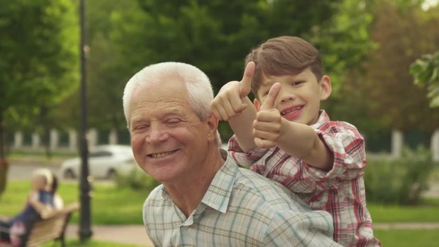 Little caucasian boy showing his thumbs on his grandpa's back. Handsome brunette boy approving spending time with his grandfather. Attractive senior man and his grandson posing for the camera in the