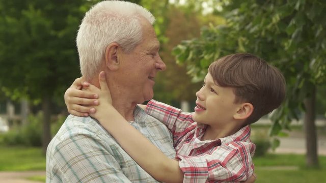 Attractive little boy holding his hands behind his grandpa's neck against background of green trees and grass. Slow motion of senior gray man holding his grandson in his arms. Close up of brunette