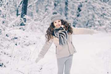 Fototapeta na wymiar Cheerful little girl in a gray jacket with a knitted hat and scarf is playing in the winter forest. Children play outdoors in a snow-covered forest. Children catch snow flakes.