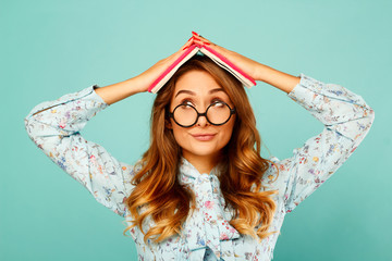 Beautiful young student holding book on her head and wearing glasses over blue background