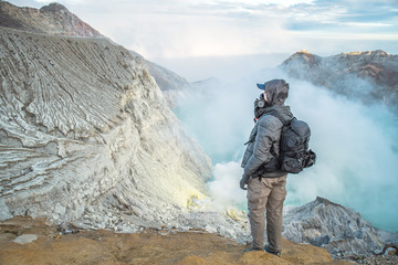 Male photographer with backpack standing on top of a mountain and enjoying sunrise, Colorful Kawah Ijen volcano