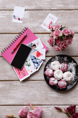 Festive background of greeting cards. Colorful zephyrs and pink notebook, crimson roses and smartphone on wooden backdrop as congratulation and corresponding concept, top view