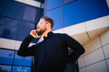 Portrait of handsome bearded businessman talking phone standing near office