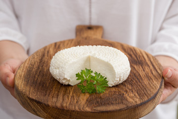 Closeup of round white homemade cheese - traditional milk creamy dairy product served with herbs on vintage wooden board in man's hands. Rustic style.