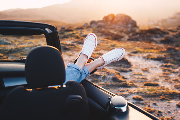 Happy woman traveler rests and admire sunset in mountains from convertible car. Woman pushes her shoes out of convertible to enjoy view and sunrise
