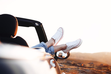 Young happy woman hipster pushes her shoes out of convertible car and admires or enjoy the view in...