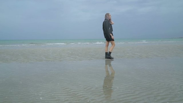 Beautiful woman walks along the beach, cold rainy weather, autumn. Sexy girl hipster or punk in a black leather jacket is walking through puddles near the sea or ocean, looks in the water reflection