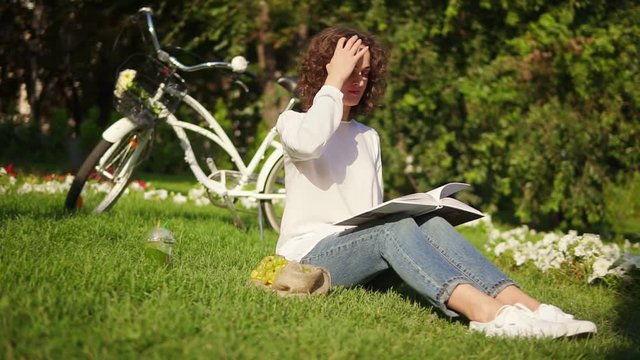 Attractive woman in white shirt and blue jeans is reading a book sitting on the grass in park during sunny warm day. Her city bicycle with basket and flowers is standing behind