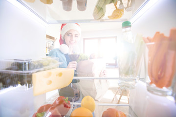 Portrait of female in santa hat standing near open fridge full of healthy food, vegetables and fruits