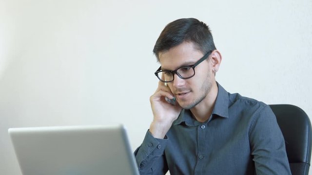 Close-up portrait of a young man wearing glasses sitting in his office in front of a monitor - working on a computer and talking on the phone. People stock footage shot. 
