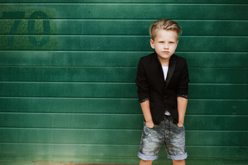 Fashionable child in the summer in the city against the background of a green wall. Trendy boy standing on the street at sunset.