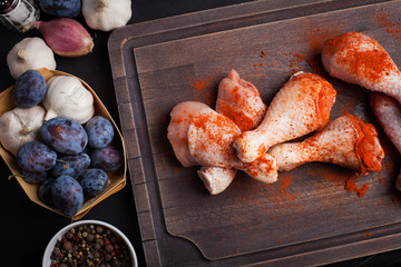 Closeup of raw chicken thighs with garlic, paprika, salt and pepper mixture on a dark background. Top view. Around the cutting Board also shallots and plums