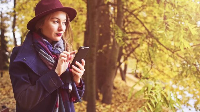 Young Woman Using SmartPhone in Autumn Park. SLOW MOTION. Girl dressed in wool coat and hat enjoying cool fall weather outdoors, texting and taking photos on smart phone. Golden falling leaves