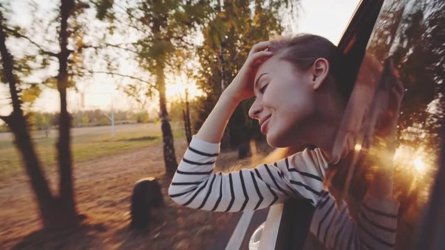 Young Woman looking out the moving Car Window, back passengers seat, 4K. Sunny Autumn Morning or Evening, Lens Flare. Happy Girl Leaning Out The Vehicle Window. Freedom and Youth. 