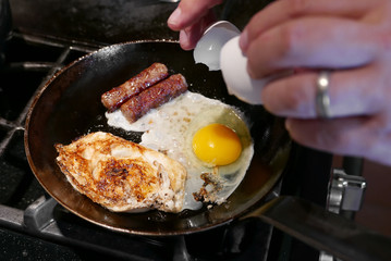 Männliche Hand schlägt ein Ei in eine gusseiserne Pfanne, in der sich bereits ein Spiegelei und zwei Würstchen befinden