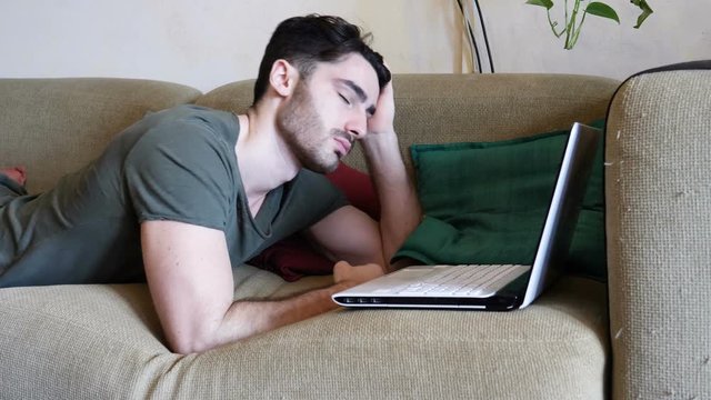 Close Up of Tired Young Man with Dark Hair Falling Asleep while Working Laptop Computer on a Couch