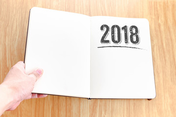 Hand holding open notebook with 2018 new year lay it on wooden table,Template mock up for adding your text.