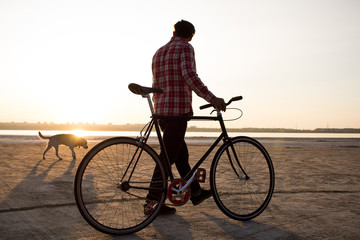 male bicycle rider walking on the sand, sunrise and lake or river background  