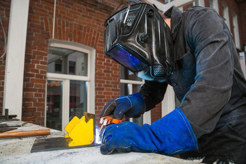 А man welder in a black work jumpsuit in a black welding mask and blue protective mitts brews an  metal  arc welding machine in the street on a summer day, in the background a bumpy old wall