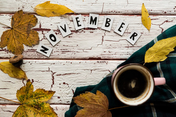 Above view of incognito woman in green checked shirt holding cup of tea or coffee on old white background, with letters in worlds " November " yellow leaves around.Home made decor for autumn season.
