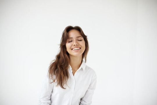 Portrait of happy joyful young woman office worker in formal blouse posing isolated at white wall with eyes closed in great pleasure and delight. Genuine positive emotions, happiness and joy