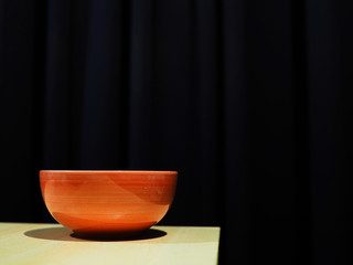 Red orange ceramic bowl with shadow on wooden table corner, black curtain background
