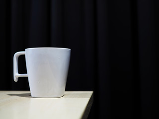 White ceramic cup mug on wooden table corner, black curtain background
