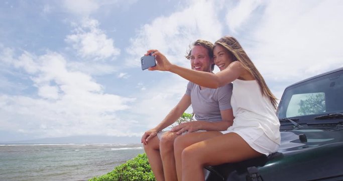 Happy couple taking selfie using smartphone at Shipwreck beach, Lanai. Young tourists are sitting on offroad vehicle trunk enjoying summer vacation on Lanai, Hawaii, taking phone pictures.