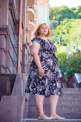 Obraz na płótnie Canvas A big cute lady in a black dress with excess weight walks in the center of the city, waiting for friends. Life of people plus size without complexes. Beautiful and pretty large woman