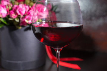 Close up of a glass with wine near stylish bouquet of pink and red roses and red ribbon in a circular black box
