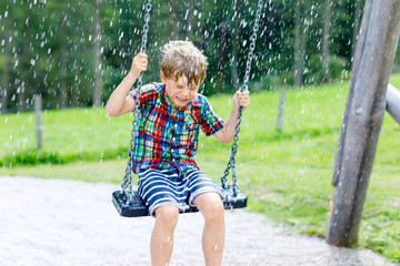 Funny kid boy having fun with chain swing on outdoor playground during rain